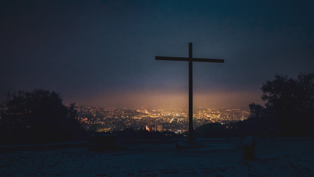 A silhouette of a cross at sunset with the lights of a city in a valley beyond